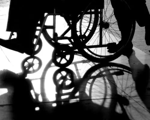 Silhouette of a person in a wheelchair being pushed, with strong shadows on the ground.