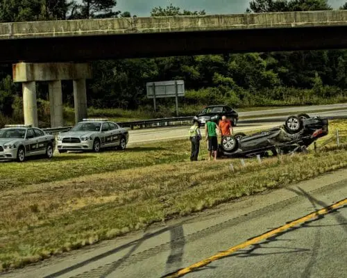 Three police cars and people stand near an overturned car beside a highway under an overpass.