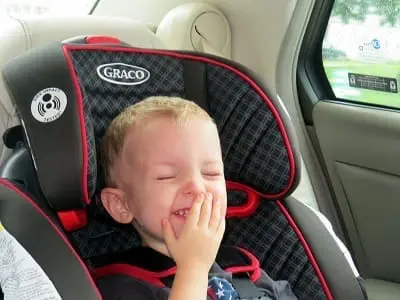 Smiling young child in a car seat covers their mouth, eyes closed, inside a car.