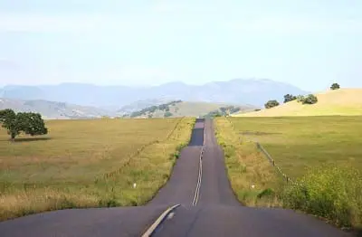 A rural road stretches through grassy hills under a blue sky with distant mountains in the background.