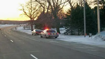A police car with flashing lights has pulled over another car on a snowy roadside at sunset.