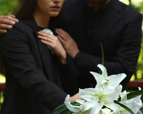 Two people dressed in black stand close together at a funeral, comforting each other beside a bouquet of white lilies, symbolizing grief and wrongful death claims in Oklahoma City.