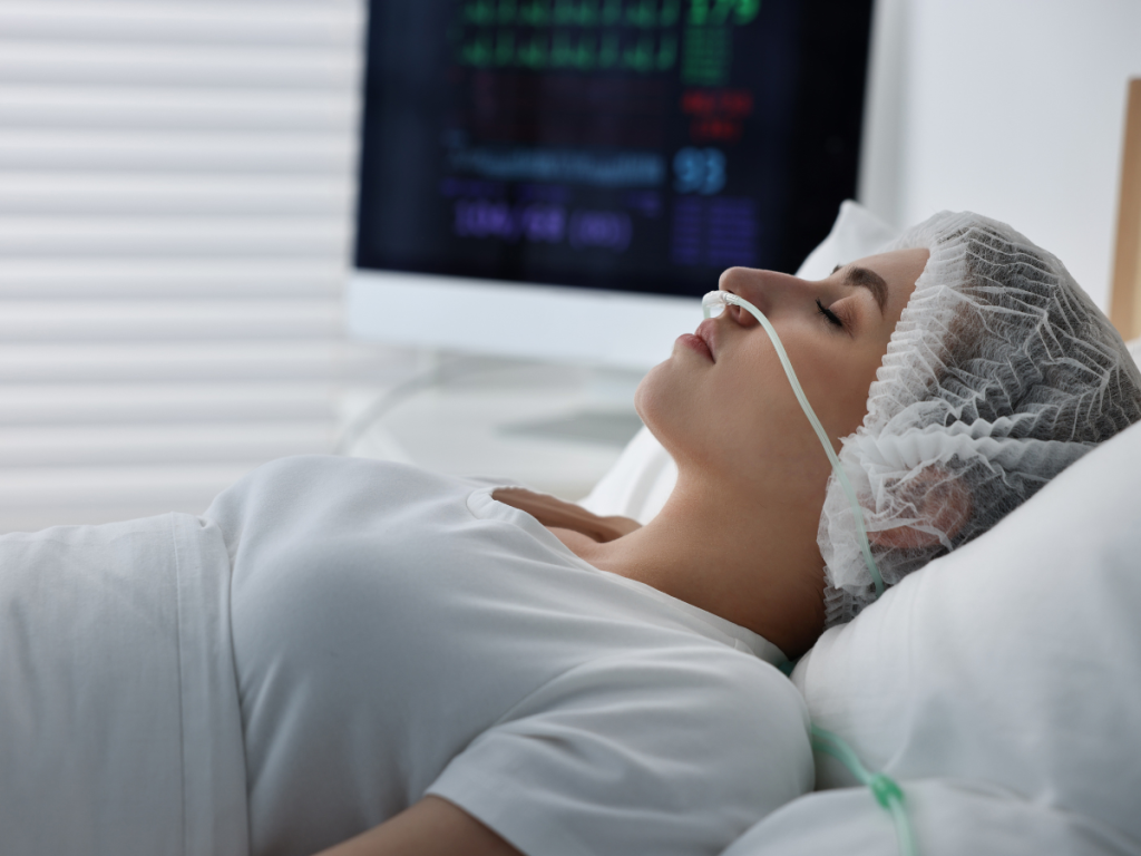 A woman lies unconscious in a hospital bed with a nasal oxygen tube and medical monitors in the background, representing the long-term impact of catastrophic brain injuries in Oklahoma City.
