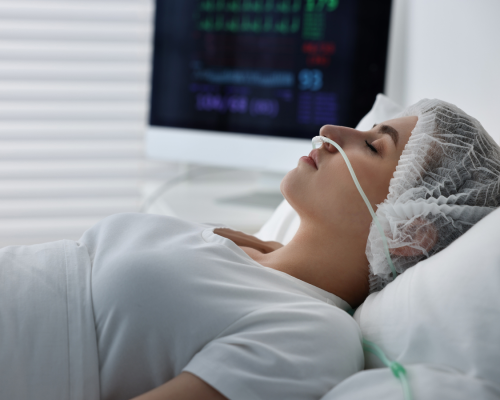 A woman lies unconscious in a hospital bed with a nasal oxygen tube and medical monitors in the background, representing the long-term impact of catastrophic brain injuries in Oklahoma City.