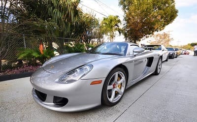 A silver Porsche Carrera GT parked on a street with several other cars in the background.